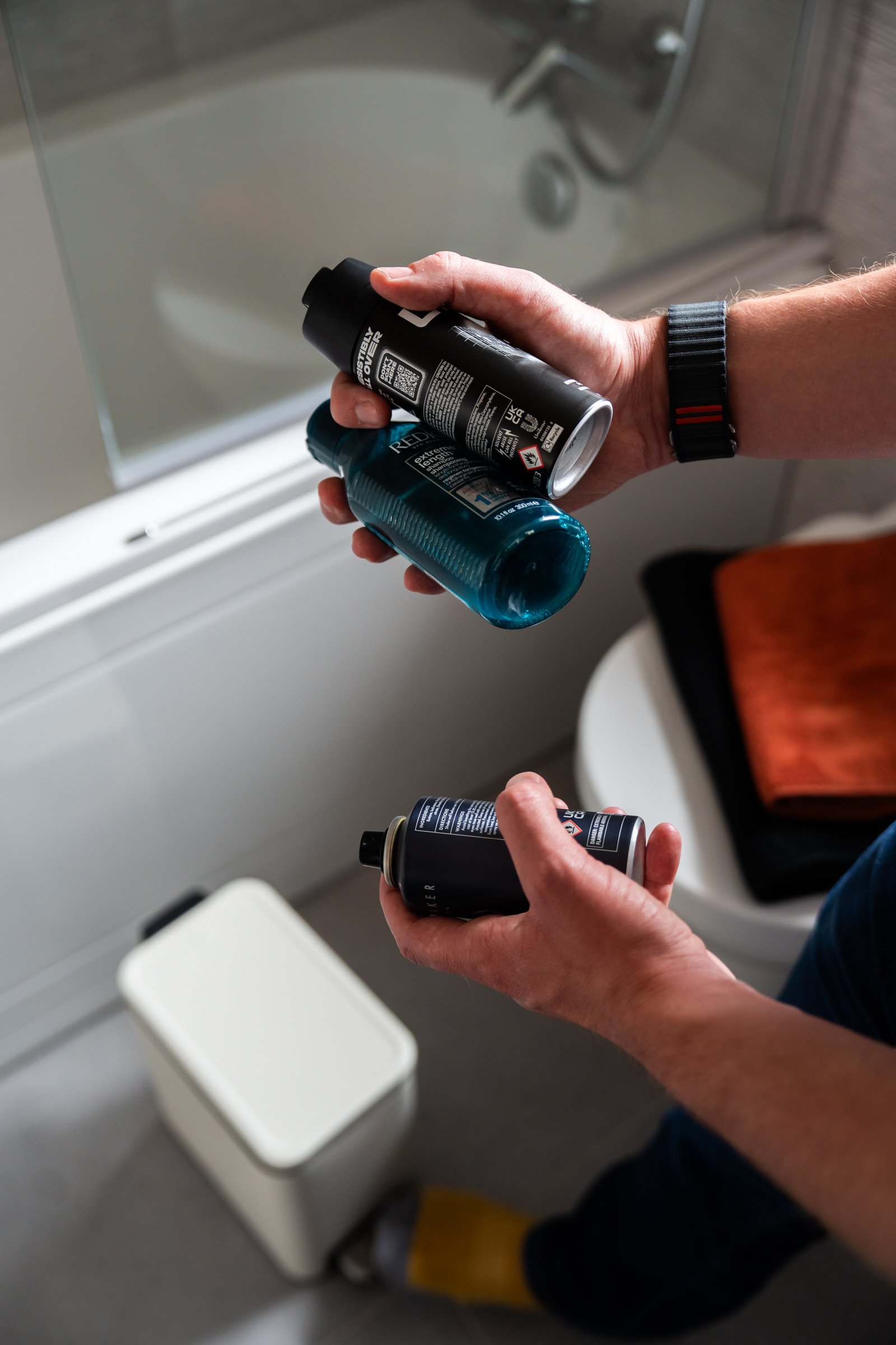 Hands hold two aerosol cans and a toiletry bottle beside a bathroom bin.