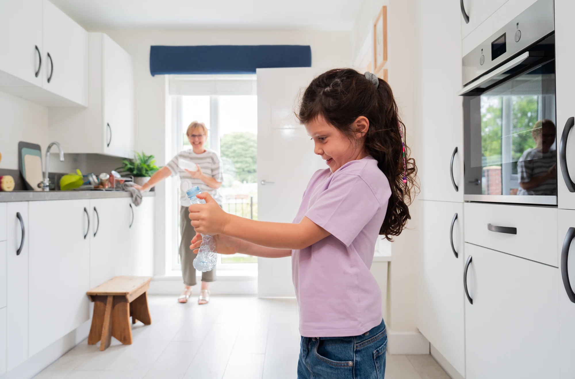A child squeezes a clear plastic bottle in the kitchen, with an adult by the sink in the background.