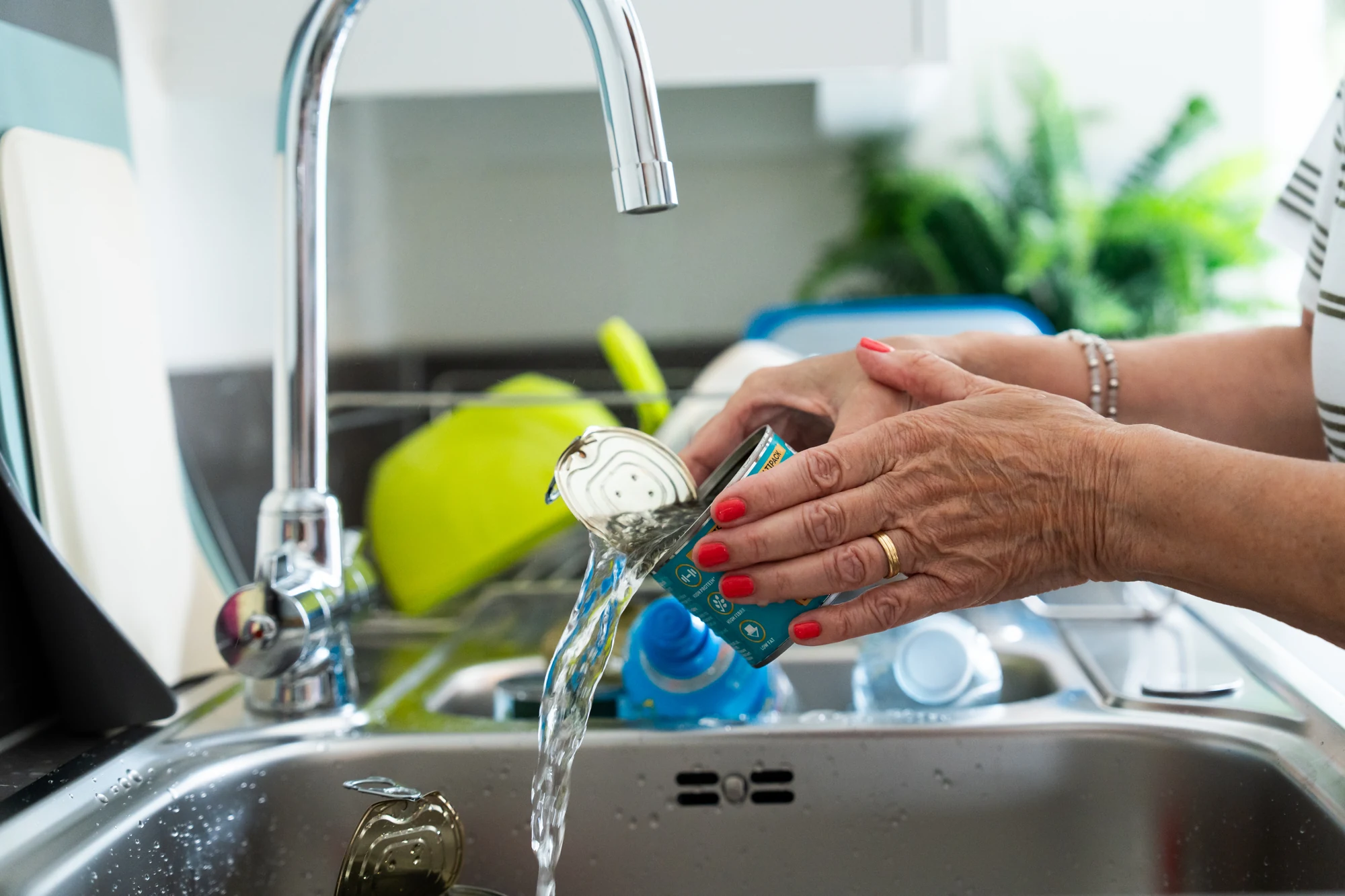 Hands tip water out of an open food tin over a kitchen sink, with washing-up beside it.